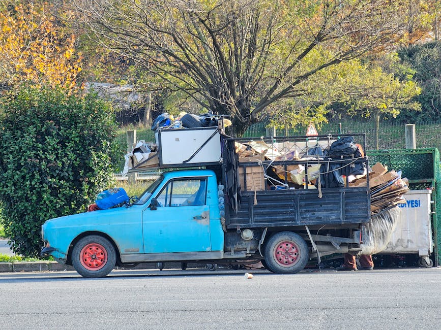 Side view of a small, blue flatbed truck parked on a street with a large pile of household items and waste materials loaded onto the cargo area. The truck is filled with cardboard boxes, wooden furniture, plastic-wrapped items, and fabric coverings, indicating preparation for a home relocation or furniture transport. Some objects, such as a white appliance and black bags, are secured with ropes, while others are loosely stacked. The vehicle's red wheels and black metal side panels contrast with the blue cab. In the background, a leafy tree with yellowing foliage suggests autumn, and a green fence borders the area. This loading process appears to be part of a professional removal service, supported by the presence of a waste disposal container and the company name Man with Van Merton, specializing in house removals and bulky-item pickups.