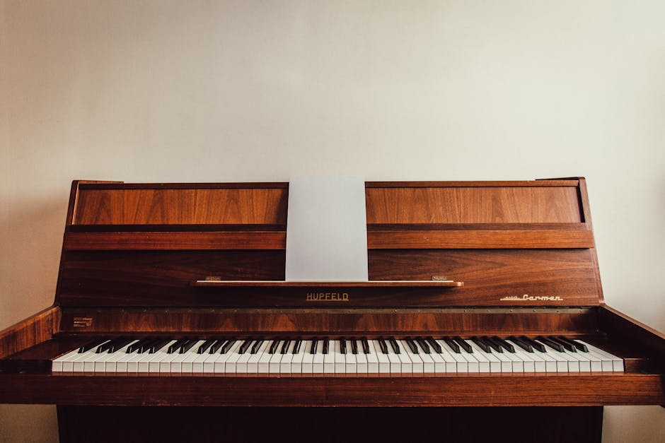 A vintage upright piano made of dark polished wood, positioned against a plain off-white wall indoors. The piano's lid is open, revealing a keyboard with black and white keys, and a music stand with a single sheet of white paper placed upright. The brand name 'Hupfeld' is visible on the front panel, alongside the model name 'Carmen.' The environment appears well-lit, with ambient lighting highlighting the wood grain and craftsmanship of the instrument. This image exemplifies furniture items typically involved in a house relocation or furniture transport process, which Man with Van Merton may handle as part of their removals services, including lifting, loading, and packing procedures for such valuable or bulky items.