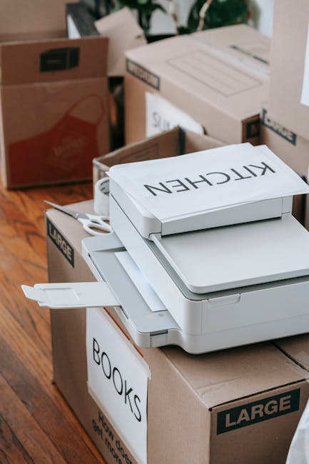 Indoor scene showing a collection of packed moving boxes and labeled cardboard containers, including a large box with a white paper label reading 'BOOKS' and another with 'KITCHEN', along with a white flatbed scanner or printer placed on top of a cardboard box. The boxes are situated on a wooden floor near a doorway or entrance to a room, with some boxes partially open revealing contents or packing materials. The environment appears to be a residential space in preparation for a home relocation, and the scene is associated with packing and moving activities carried out by Man with Van Merton as part of their removals service in Colliers Wood, Merton.