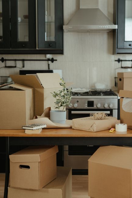 Inside a modern kitchen with light-colored tiled walls and dark upper cabinets, there is a wooden table cluttered with packing supplies such as cardboard boxes, some opened and sealed with tape, a small potted plant, and wrapping materials. Behind the table, a stainless steel oven and stovetop are visible, with a range hood above. On the right side, a cardboard box is partially open, and a packing roll is placed nearby. The scene suggests a home relocation or decluttering process, possibly organized by Man with Van Merton as part of their removals service, preparing items for transportation. The overall environment is well-lit, clean, and ready for a furniture transport or packing and moving activity.