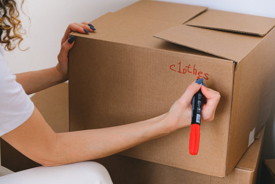 Indoor scene showing a collection of packed moving boxes and labeled cardboard containers, including a large box with a white paper label reading 'BOOKS' and another with 'KITCHEN', along with a white flatbed scanner or printer placed on top of a cardboard box. The boxes are situated on a wooden floor near a doorway or entrance to a room, with some boxes partially open revealing contents or packing materials. The environment appears to be a residential space in preparation for a home relocation, and the scene is associated with packing and moving activities carried out by Man with Van Merton as part of their removals service in Colliers Wood, Merton.