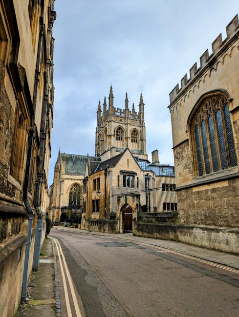 A narrow street in a historic city area with stone buildings and a Gothic-style church featuring tall spires and large arched windows in the background. The street is paved with asphalt and has a double yellow line along the curb. On either side, there are aged stone facades with detailed architectural features, including a mix of small window openings and decorative stonework. The scene appears calm and residential, with no vehicles or pedestrians visible. This setting reflects a typical environment for house removals or home relocation services, where careful planning for the transport of furniture and packing materials is essential. Man with Van Merton’s removals team may operate in such areas, ensuring efficient and secure furniture transport within historic urban locations, supporting local house moves, and adhering to local disposal regulations after a move.
