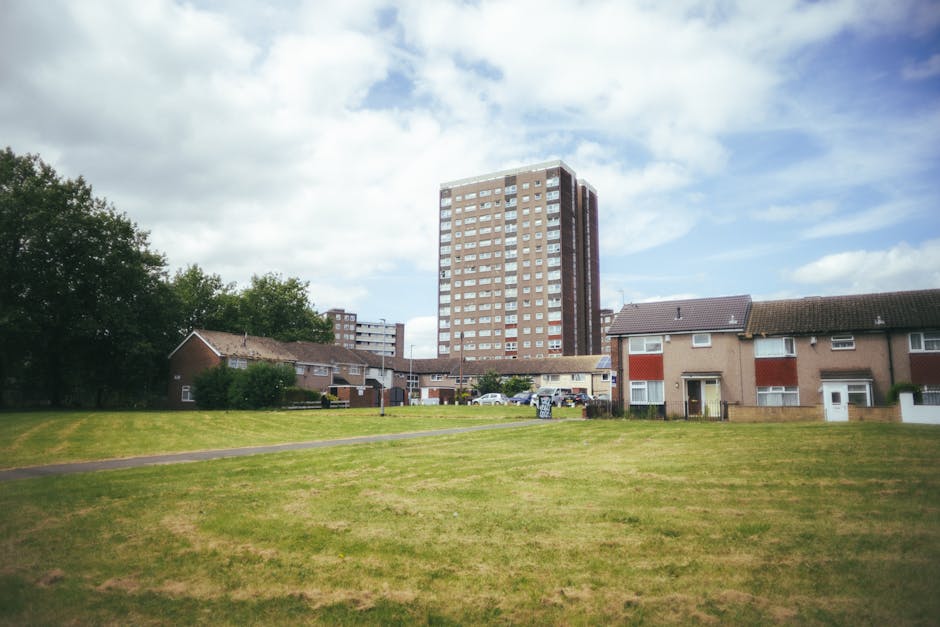 A view of a residential area with a grassy common space in the foreground, featuring a paved pathway running through the middle. In the background, there are several low-rise brick houses with pitched roofs, some of which have front gardens and small fences. A tall, multi-storey high-rise building made of brick and concrete stands prominently behind the houses, with multiple windows and balconies. The scene is captured during daylight with partly cloudy skies and natural lighting. Although no furniture or boxes are visibly being moved, the setting suggests a typical home relocation environment often managed by removals companies such as Man with Van Merton, with space suitable for furniture transport and loading process activities.