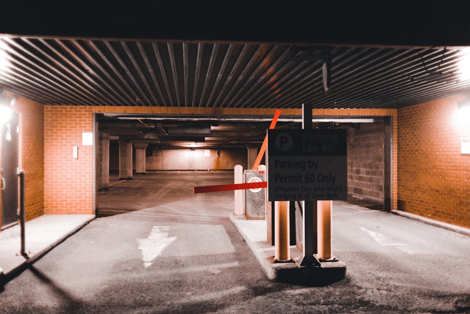 The image depicts the entrance to an underground parking garage within a residential or commercial building. The garage features a brick wall on the left side and dark horizontal ceiling panels above, with artificial lighting illuminating the space. A metal barrier arm, painted in red and white, is lowered across the entry point, mounted on columns with reflective safety strips. A parking sign is visible behind the barrier, indicating parking regulations, including a permit requirement for parking lot 60. The pavement leading into the garage is marked with worn white directional arrows and lines, guiding vehicles inward for home relocation or furniture transport during the moving process. The interior of the garage extends into a dimly lit area, with concrete pillars supporting the structure, suggesting it is prepared for accommodating move-related vehicle loading or unloading activities. This setting aligns with relocation services offered by Man with Van Merton, illustrating logistics involved in house removals and packing movements.