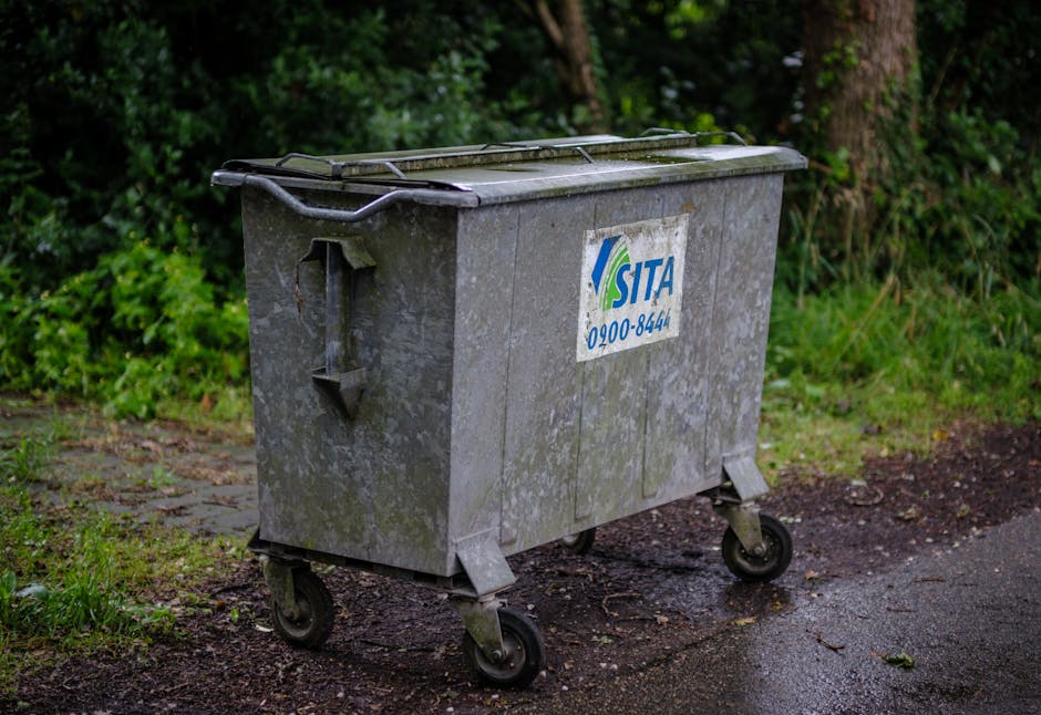 A large, weathered metal wheeled dumpster positioned on a paved area beside a patch of grass and greenery, with a background of trees and bushes. The dumpster features a partially open lid, metal handles, and a blue and white label with the logo and contact number for SITA. The scene suggests an outdoor waste collection site or preparation for removal, likely related to a home relocation or clearance process. There are no people visible, and the surroundings are natural with soft lighting, emphasizing the utility and placement of the waste container in the context of moving and clearance services, such as those offered by Man with Van Merton, especially in relation to local permit arrangements like skip permits and parking suspensions in Merton.