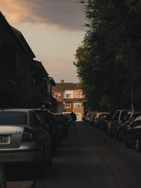 A narrow residential street in Morden SM4 during early evening with parked cars lining both sides of the paved roadway. On the left, there are multi-storey brick and timber apartment buildings with balconies, while on the right, tall leafy trees provide shade. In the distance, a person wearing dark clothing is seen walking along the street near a white garage door. The scene is illuminated by soft, fading natural light from the sunset. This urban environment depicts a typical setting for home relocation or furniture transport within a neighbourhood, which Man with Van Merton specializes in, including parking management and logistics for house removals and packing during moving day.