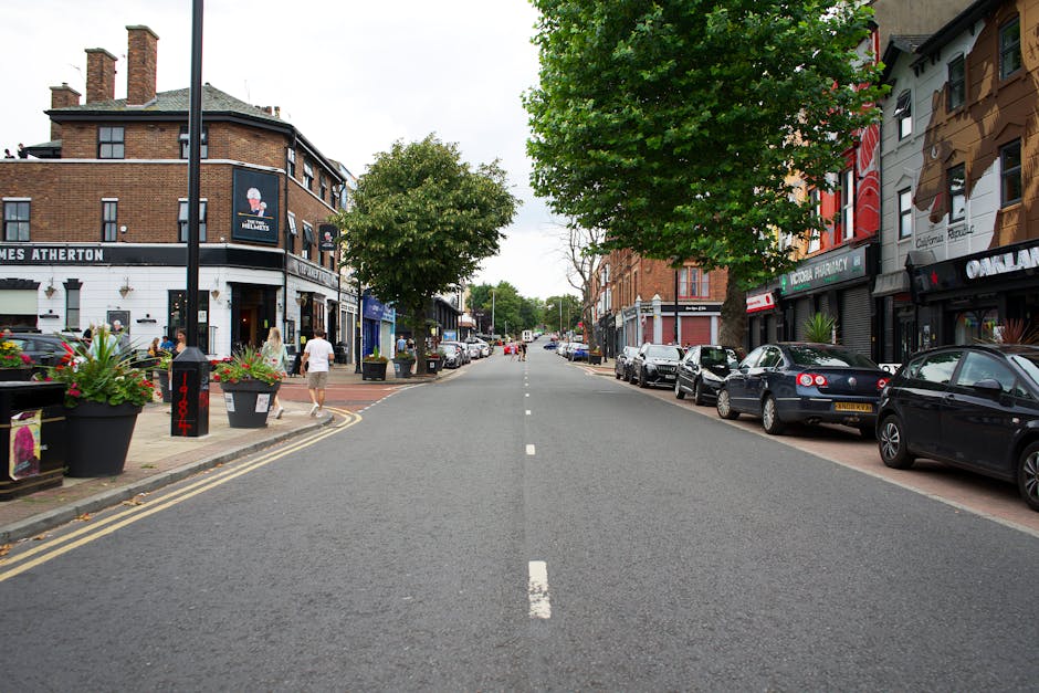 A wide view of Trinity Road in Mitcham showing a street lined with two and three-storey commercial buildings and residential properties. Parked cars of various models and colors are aligned along both sides of the road on the pavement. Several pedestrians and shoppers are walking along the sidewalk, some near a furniture shop and cafes. The street features greenery, including a large leafy tree on the right side, with planters containing flowers placed along the sidewalk on the left. The scene is well-lit with natural daylight, and the overall environment appears calm and typical of a busy urban area. This setting illustrates the urban context where house removals and furniture transport services by Man with Van Merton could facilitate home relocation or moving logistics in this area, with possible loading zones or vehicle access nearby.