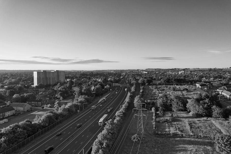 Black and white aerial photograph of a residential area depicting a multi-lane motorway with several vehicles, including cars and a bus, running through the suburb. Surrounding the motorway are rows of houses, some with gardens, and scattered trees. To the right side of the image, there's an open patch of land with a few small structures and what appears to be farmland or undeveloped space. In the background, taller apartment buildings and distant hills are visible under a partly cloudy sky. The scene captures an urban environment with clear evidence of home relocation or transportation activity, with the motorway and nearby properties suggesting the context of house removals and moving logistics supported by [COMPANY_NAME], incorporating the process of furniture transport and packing during a home move.