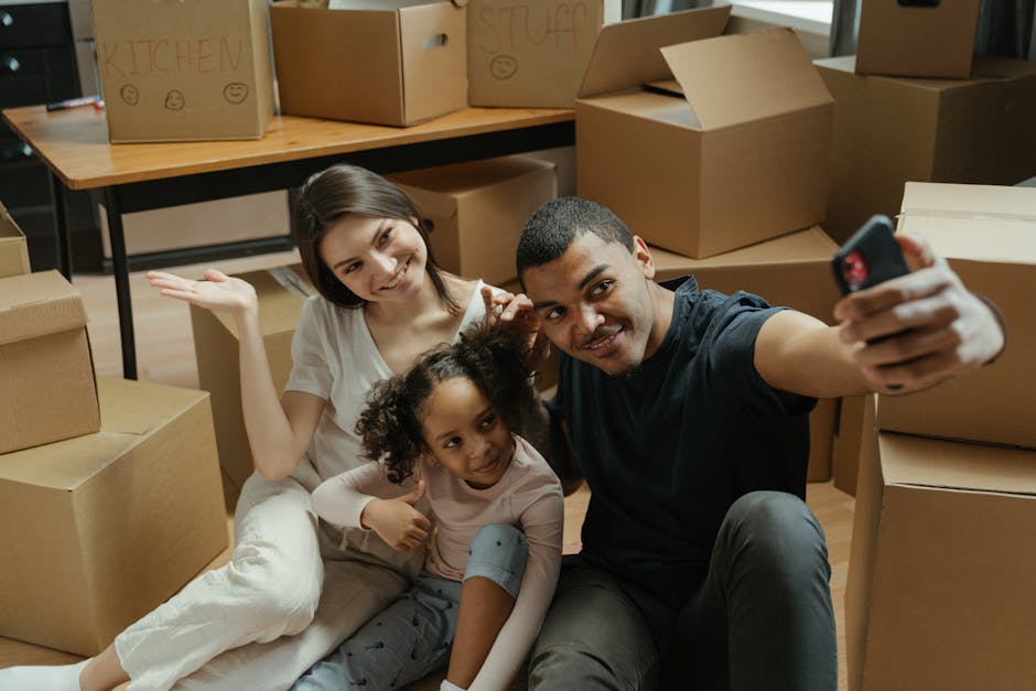A family of three, including a woman with shoulder-length brown hair, a young girl with curly hair wearing a light pink top and grey pants, and a man with short dark hair and a black T-shirt, seated on the floor surrounded by numerous packed cardboard boxes with handwritten labels such as 'KITCHEN' and 'STUFF', inside a room equipped with a wooden table and metal legs. The family members are smiling and taking a selfie with a smartphone, capturing a moment during their house move. The background shows the boxes stacked near the doorway, indicating an active packing process as part of a house relocation. The boxes are of various sizes, some open and sealed with packing tape, some displaying printed or handwritten labels. The scene suggests a busy moving day with furniture and belongings prepared for transport, aligned with professional moving services offered by Man with Van Merton for urgent same-day relocations.
