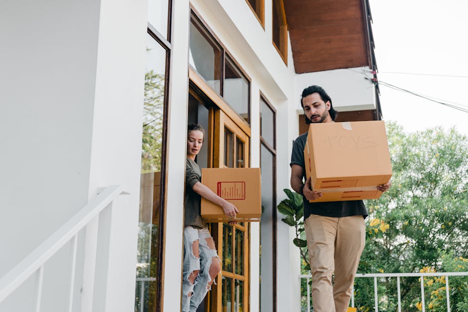 A man and a woman are standing on the porch of a modern residential building, each holding a cardboard box filled with household items. The woman, wearing a casual grey jacket and ripped jeans, is reaching towards the doorway, while the man, dressed in a dark shirt and beige trousers, is carrying a box labeled 'TOYS'. The porch is adjacent to large glass windows and a white exterior wall, with a potted plant visible near the doorway. The scene takes place during daylight, with green trees and an outdoor setting in the background. The individuals are involved in a home relocation process, preparing to load boxes into a vehicle as part of a professional removals service by Man with Van Merton, focusing on efficient packing and moving logistics during a house move.