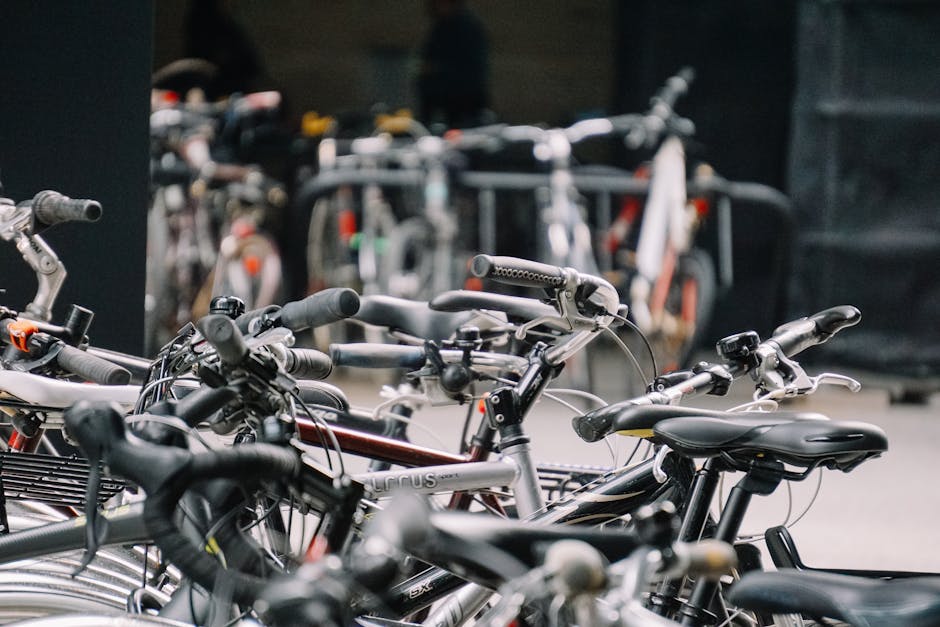 A close-up view of multiple bicycles parked closely together on a paved surface, with some bikes stacked and secured with locks; in the background, there's a black storage unit and a row of more bicycles, indicating an urban or residential setting. The bikes vary in colour and design, including silver, black, and red frames, with different handlebar styles and seating. The image captures the scene during daytime with natural lighting, typical of a bicycle storage area outside a property, suitable for home relocation or moving services like those offered by Man with Van Merton, which specialises in removals and furniture transport, including loading and packing logistics.