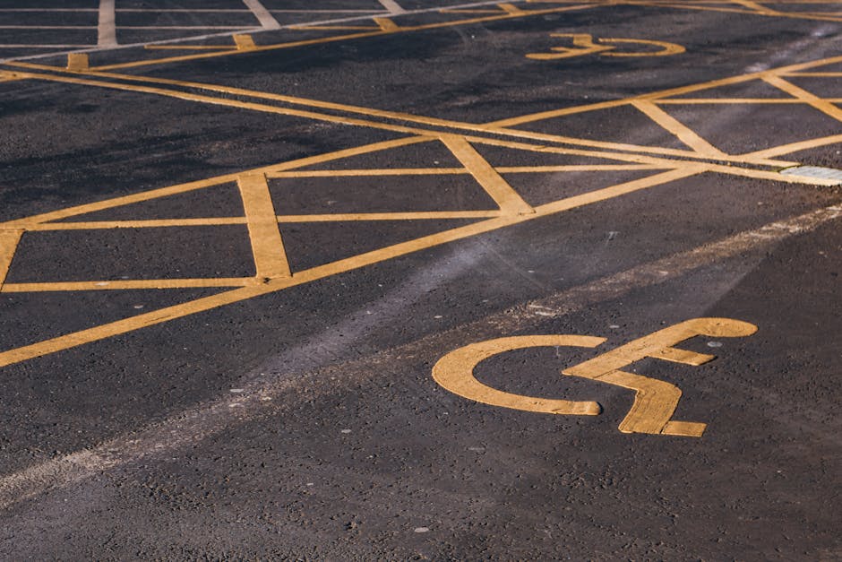 Close-up view of an outdoor parking area with a designated accessible parking space marked by bright yellow paint, including a wheelchair symbol on dark asphalt. Surrounding the space are yellow hatch markings creating a border or no-parking zone. The surface shows signs of wear with some uneven patches and slight fading of the paint. This scene is part of a typical parking lot used for home relocation or furniture transport services, with no vehicles present in the image. The parking area is likely used for loading or unloading household items, with clear access for removal vans operated by companies such as Man with Van Merton. The image captures the environment where residential moving logistics might be coordinated, supporting the accessibility requirements and efficient drop-offs during a move from Wimbledon Common, with attention to parking regulations and space planning for house removals.